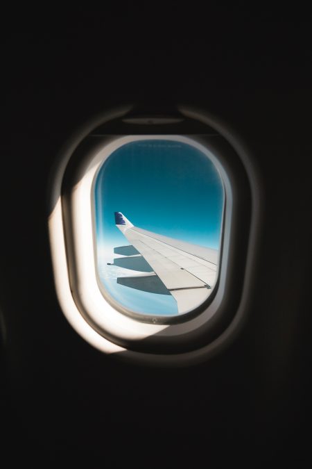 Airplane window looking outside to a blue sky with white clouds and plane wing.
