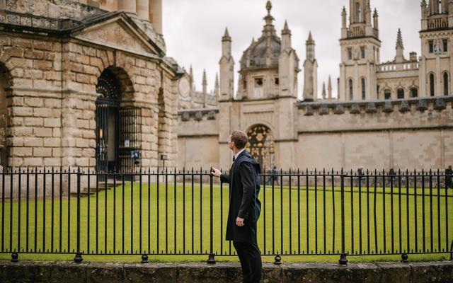 Jack Replinger stood in black traditional Oxford robes, by railings looking at the Oxford Radcliffe Camera