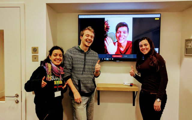 three students standing around a tv screen with a student on a virtual call.