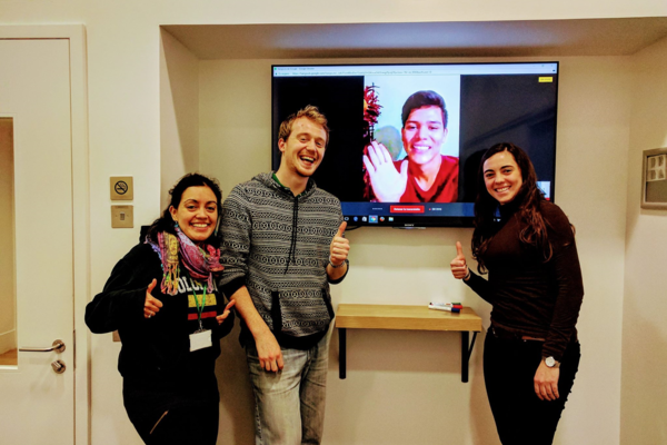 three students standing around a tv screen with a student on a virtual call.
